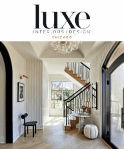 A modern entryway with a black and white color scheme, featuring a marble table, a black chair, and a white pouf. The staircase has black railings and wooden steps, leading to a bright and airy space with large windows.